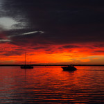 A sailboat sits anchored in the distance with a sky exploding in orange and red like a hovering fire as the sun sets on the horizon. The fierce oranges and reds reflect off of the gentle waves of the ocean.
