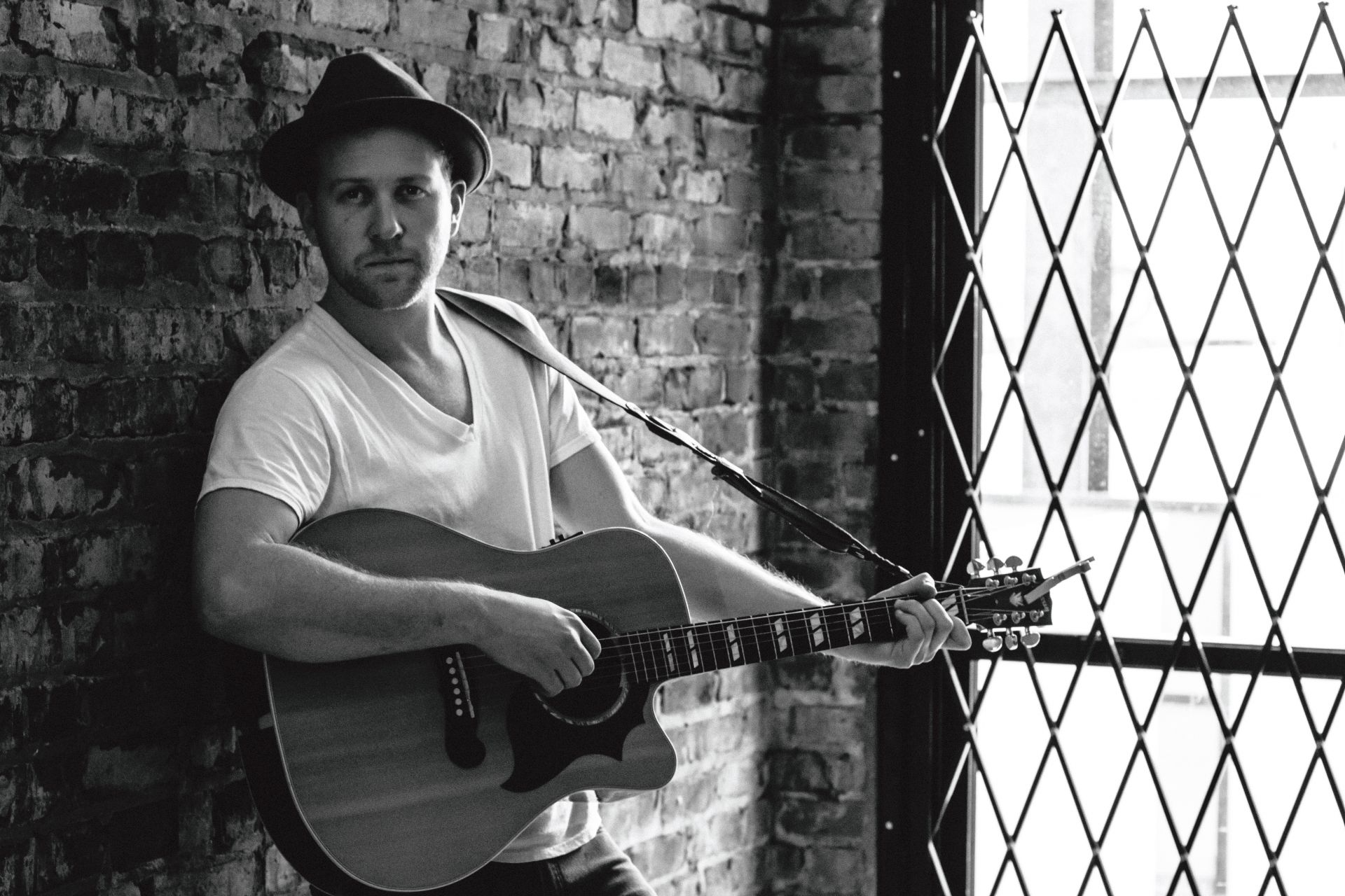 Portrait photo of Cody Marlowe leaning against a brick wall playing an acoustic guitar