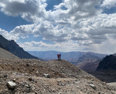 Lonnie Bedwell and Steve Baskis stand far in the distance with fluffy clouds above them overlooking the mountain ridgeline near Aconcagua.